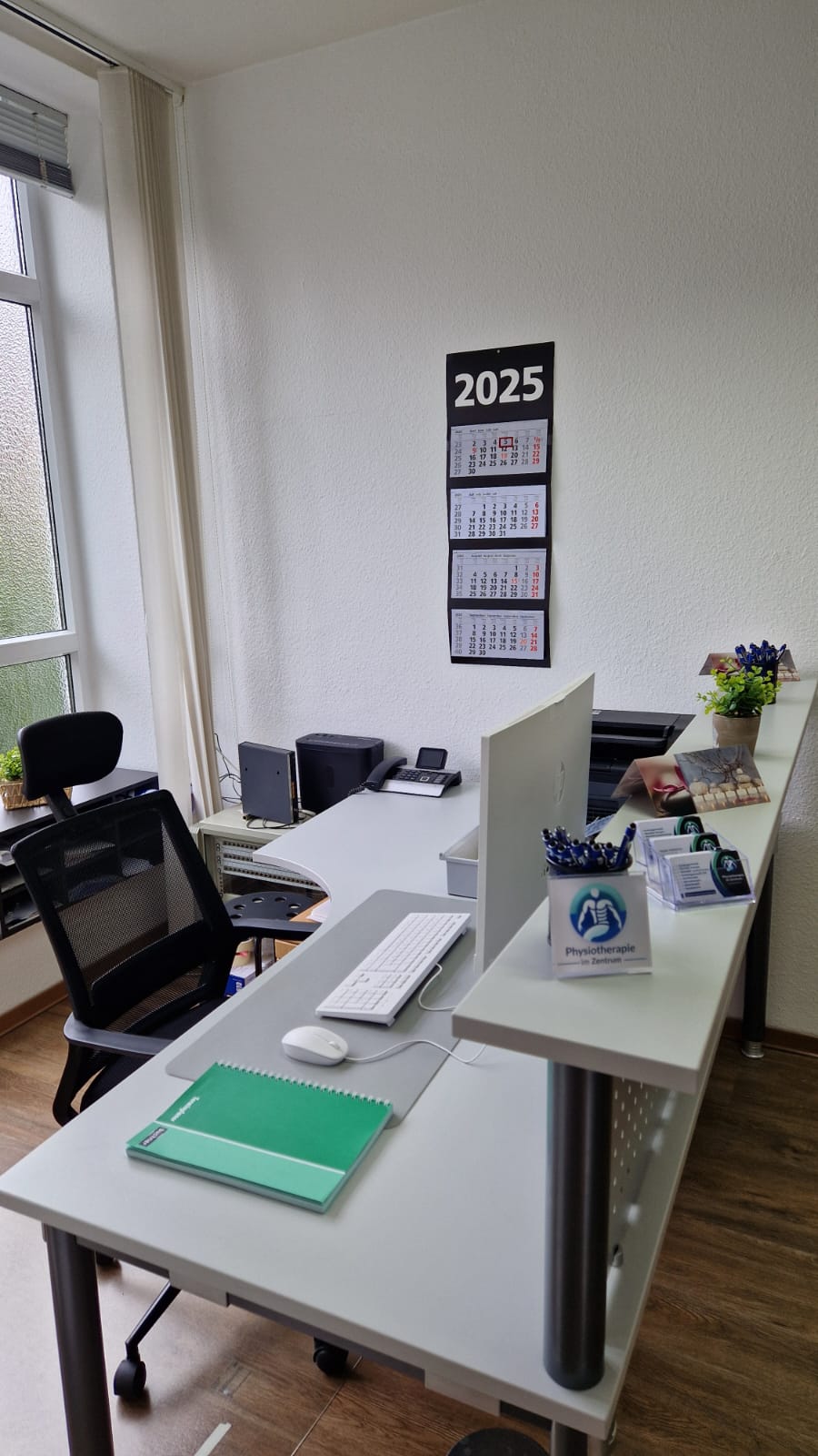 Empfangsbereich der Praxis mit Schreibtisch, Kalender und Büroausstattung.