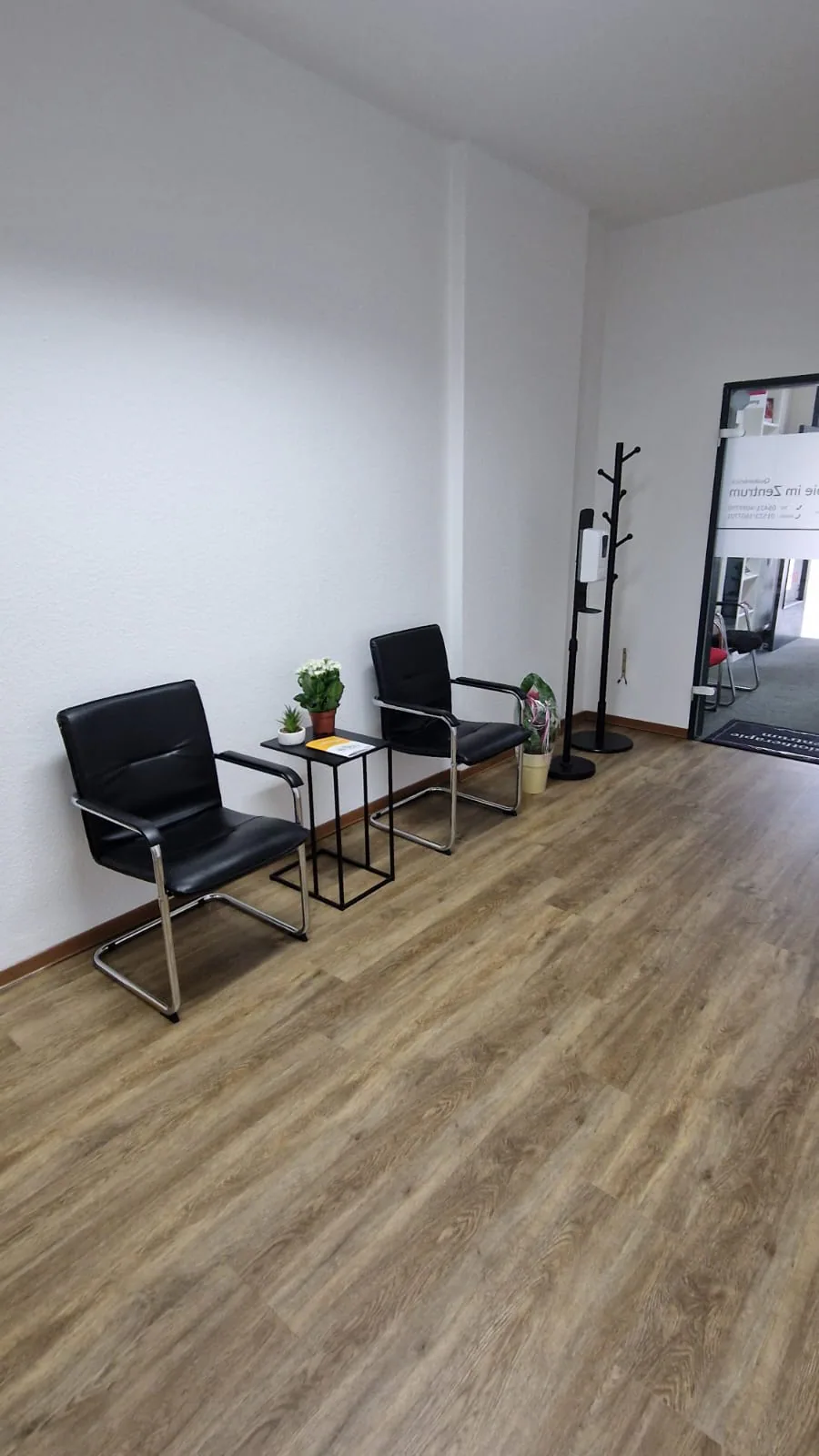 Modernes Wartezimmer mit schwarzen Sesseln, Pflanzen und Desinfektionsspender.